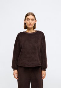 Brauns fleeces džemperis ar apaļu kaklu, garām piedurknēm un brīvu piegriezumu. Ievērojama mīksta tekstūra un ribotas piedurknes.