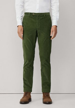 Pantalon en velours côtelé vert avec une coupe slim, se ferme par un bouton, avec des passants de ceinture et un tissu texturé. Porté avec des bottes en cuir marron à lacets.