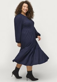 Vestido azul marino de manga larga con tejido texturizado, falda de volantes y silueta holgada. Llevado con botas negras hasta el tobillo, con punta afilada.