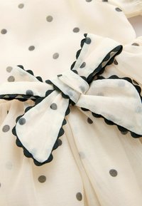 Jasnobeżowa tkanina z czarnymi groszkami i dużą kokardą. Kokarda ma falbaniaste krawędzie w czerni, co dodaje tekstury do wzoru.