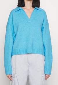 Hellblauer Strickpullover mit V-Ausschnitt und einem gerippten Saum. Verfügt über einen klassischen Kragen und eine lockere Passform. Kombiniert mit hellen Cargohosen.