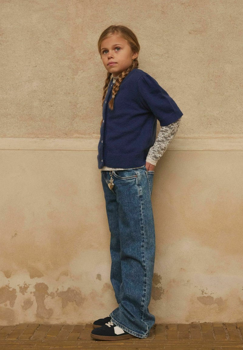 Ragazzina con i capelli intrecciati, che indossa un cardigan blu navy su una maglietta in pizzo a maniche lunghe, jeans blu e scarpe nere, in piedi contro un muro beige.