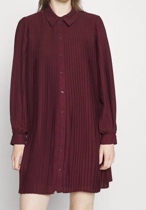 Kvinde iført en burgunderfarvet, plisseret skjortekjole med lange ærmer og knapper, samt spids krave, mod en ensfarvet baggrund.