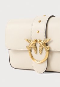 Κρέμα δερμάτινη τσάντα χειρός με λεία υφή, διαθέτοντας χρυσές λεπτομέρειες σε σχήμα πουλιού και τρουκς στην κλειστή θήκη.