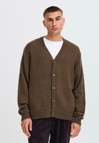 Brown knitted cardigan with a v-neck, five buttons, ribbed cuffs, and hem. Worn over a white shirt, paired with dark corduroy pants.