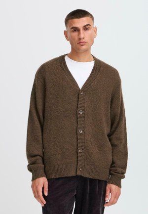 Brun strikket cardigan med v-udskæring, fem knapper, ribbekantede ærmer og kant. Båret over en hvid skjorte, parret med mørke fløjl bukser.