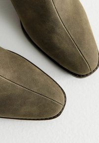 Bottines en suède vert olive avec des bouts pointus, des détails cousus et des accents de semelle foncés. Texture lisse et design élégant.