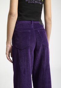 Pantalons en velours côtelé violet à jambes larges, dotés de poches arrière et d'un motif en relief texturé. La taille est maintenue par une petite étiquette noire.
