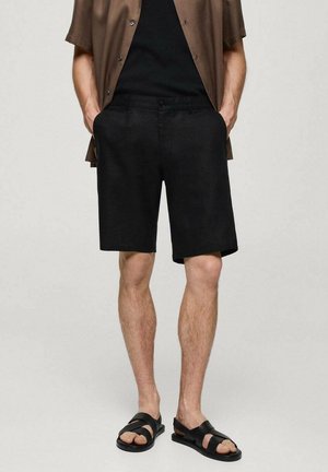 Mężczyzna w czarnych szortach, czarnej koszulce, brązowej koszuli z krótkim rękawem i czarnych sandałach stoi z rękami w kieszeniach.