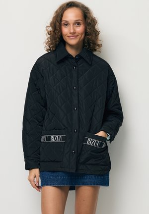 Vrouw met een zwarte doorgestikte jas met het BIZUU-logo op de voorzakken en een spijkerrok, staand tegen een effen lichte achtergrond.