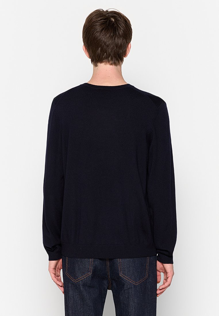 Marineblauwe sweater met lange mouwen, een ronde hals en geribbelde boorden. Gladde textuur, minimale details en een relaxte pasvorm. Gedragen met een donkere spijkerbroek.