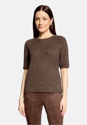 Vrouw met steil, donker haar die een bruine, zachte korte mouw trui en bijpassende bruine suède broek draagt, staand voor een effen achtergrond.