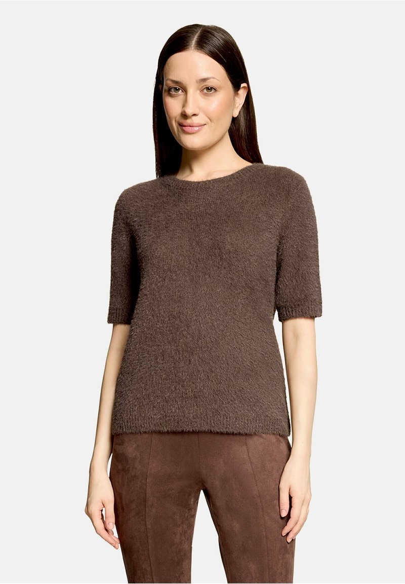 Femme aux cheveux raides et foncés portant un pull court à manches courtes marron duveteux et un pantalon en daim marron assorti, debout sur un fond uni.