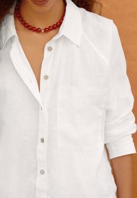 Chemise en lin blanc avec un devant à boutons, une poche poitrine unique et des manches retroussées. Elle présente des boutons gris clair et une finition texturée.