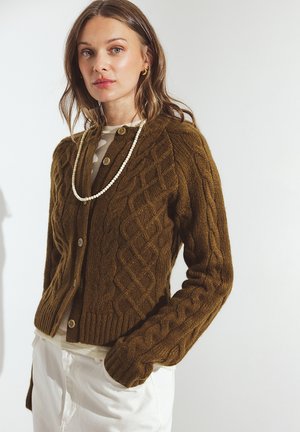 Tume oliivroheline kudumitega kardigan, millel on kaelus ümmarguse kujuga ja puidust nööbid. Kardigan on kantud heleda särgi üle ja kombineeritud valgete pükste juurde.