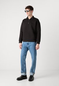 Hanorac negru cu guler cu fermoar, având un logo subtil; combinat cu blugi albaștri deschiși și pantofi negri. Modelul poartă ochelari de soare.