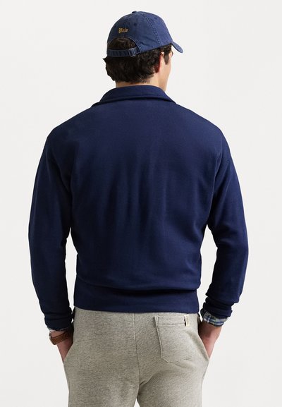 Pulover bleu marin cu o textură netedă, având culoare și mâneci lungi, asortat cu pantaloni de trening gri deschis și o șapcă bleumarin.