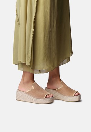 Nohy v béžových semišových otvorených platformových sandáloch na tenkých podrážkach s prešívanými okrajmi, v páre s voľne splývajúcou olivovo zelenou sukňou.