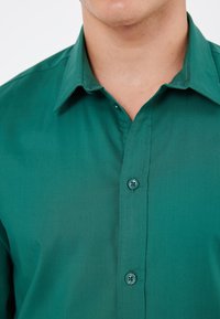 Grüne Hemd mit Knopfleiste, glatter Textur, flachem Kragen und zwei sichtbaren Knöpfen vorne. Sauberes, minimalistisches Design.