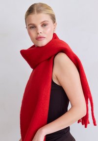 Rode sjaal van zachte, gestructureerde wol, met een brede, langwerpige vorm en rafelige uiteinden, gedrapeerd om de hals.