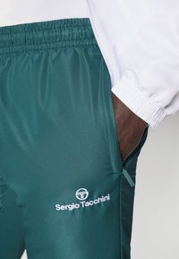 Zaļi sporta bikses, izgatavotas no teksturēta auduma, ar elastīgu jostasvietu, ar rāvējslēdzēja kabatām un balto logotipu izšūtu uz augšstilba.