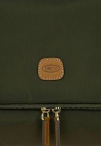 Olijfgroene stoffen tas met een gladde textuur, voorzien van een tan lederen label en twee metalen ritsen met contrasterende banden.