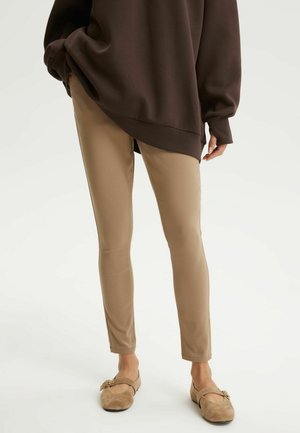 Osoba ubrana w beżowe wąskie spodnie, brązowy oversize'owy sweter oraz jasnobrązowe zamszowe baleriny Mary Jane z ozdobną klamrą, stojąca na białym tle.