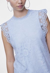 Ljust blå spetsblus med vågiga kanter och blommönster, korta ärmar och en texturerad yta. Stora silverfärgade hoopörhängen.