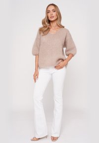 Beiger, flauschiger Pullover mit V-Ausschnitt und Ballonärmel, kombiniert mit weißen Schlagjeans. Das Model steht mit einer Hand auf der Hüfte.