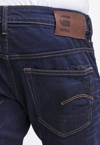 Jeans in denim blu scuro con una texture liscia, cuciture gialle e un'etichetta in pelle sulla vita che mostra "G-STAR RAW."