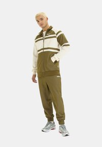 Tuta sportivo verde oliva e bianco con giacca con zip, dotata di strisce e tasche laterali, abbinata a pantaloni coordinati e sneakers grigie.