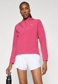 Veste légère rose avec capuche, fermeture éclair et accents réfléchissants. Associée à un short de sport blanc et une bouteille d'eau noire.