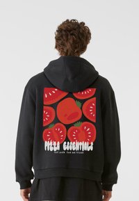 Fekete kapucnis pulóver, színes grafikával a hátsó részén, amelyen piros paradicsomok, zöld kiegészítők és a "Pizza Essentials" felirat látható fehér színben.
