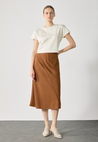 Bílý zkrácený top s krátkými rukávy, doplněný hnědou sukní po kolena s volným střihem. Modelka má na sobě světlé, texturované boty.