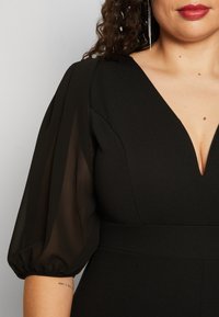 Crna haljina s dubokim V-izrezom, koja ima prozirne, napuhane rukave i oblikovani siluetu. Glatka tekstura s minimalnim detaljima.