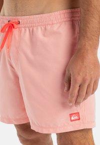 Prúžkované červené plavky s elastickým pásom a oranžovým šnúrkou. Obsahujú malú nášivku s logom na boku. Hladká textúra.