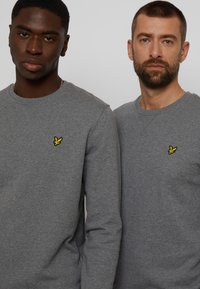 Deux hommes portant des sweatshirts en coton gris à manches longues. Chacun a un petit logo jaune brodé sur la poitrine. Texture lisse.