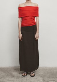 Haut rouge à épaules dénudées associé à une jupe maxi marron avec un cordon de serrage à la taille, porté avec des tongs noires sur une surface texturée.