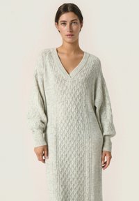 Abito in maglia leggera di colore grigio chiaro, con scollatura a V profonda, maniche lunghe a sbuffo e polsini a coste; texture morbida e vestibilità rilassata.