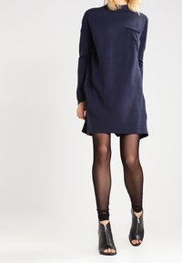 Stickad klänning i marinblått med hög hals, långa ärmar och sidslitsar. Har en bröstficka. Kombineras med genomskinliga svarta strumpbyxor och svarta öppna stövlar.