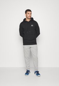 Sudadera negra con bolsillo frontal y capucha ajustable, combinada con pantalones de chándal gris claro y zapatillas deportivas azules. Diseño simple, sin patrones.