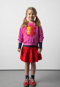 Różowa bluza z żółtymi i czerwonymi postaciami z kreskówek, zestawiona z czerwoną spódnicą o falbaniastym wykończeniu oraz czerwonymi trampkami z beżowymi akcentami.