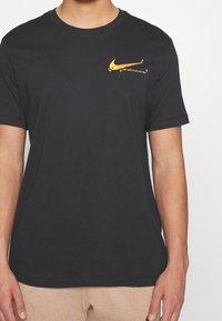 Svart t-shirt med kort ärm i bomull, med en gul Nike-logotyp och textdesign på vänster bröstområde.