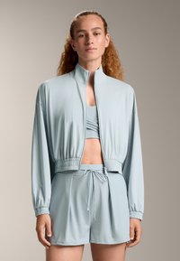 Svijetloplavi sportski set koji uključuje kratku jaknu s visokim ovratnikom, elastičnim pojasom i usklađenim šorcima svezanom žicom. Mekana i glatka tekstura.