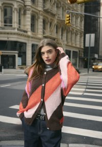 Multicolored cardigan featuring diagonal orange, pink, and brown patterns, with button detailing and a soft, textured fabric.