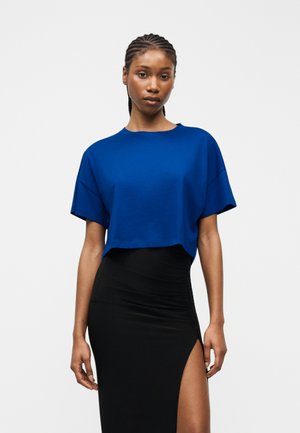 Kvinde med flettet hår iført en løs blå cropped top og sort nederdel med høj talje og slids op til låret, stående foran en ensfarvet baggrund.