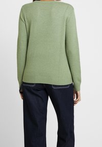 Pull en maille vert à manches longues avec ourlet côtelé, offrant une coupe décontractée. Porté avec un jean droit bleu foncé.