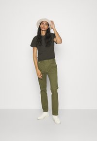 Maglietta verde scuro, pantaloni verde oliva e sneakers bianche. Indossato con un cappello beige, il modello si trova di fronte a uno sfondo bianco. Capelli lunghi visibili.