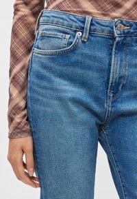 Los vaqueros de mezclilla azul presentan un diseño clásico de cinco bolsillos, cremallera y cierre de botón. La tela tiene una textura ligeramente desgastada con un sutil efecto desteñido.