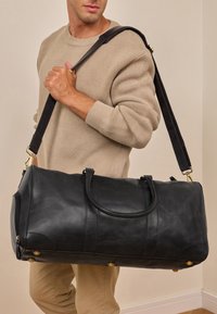 Sort læder duffel bag med to håndtag, en aftagelig skulderrem og guldsmykker. Glat tekstur med paneldetaljering.